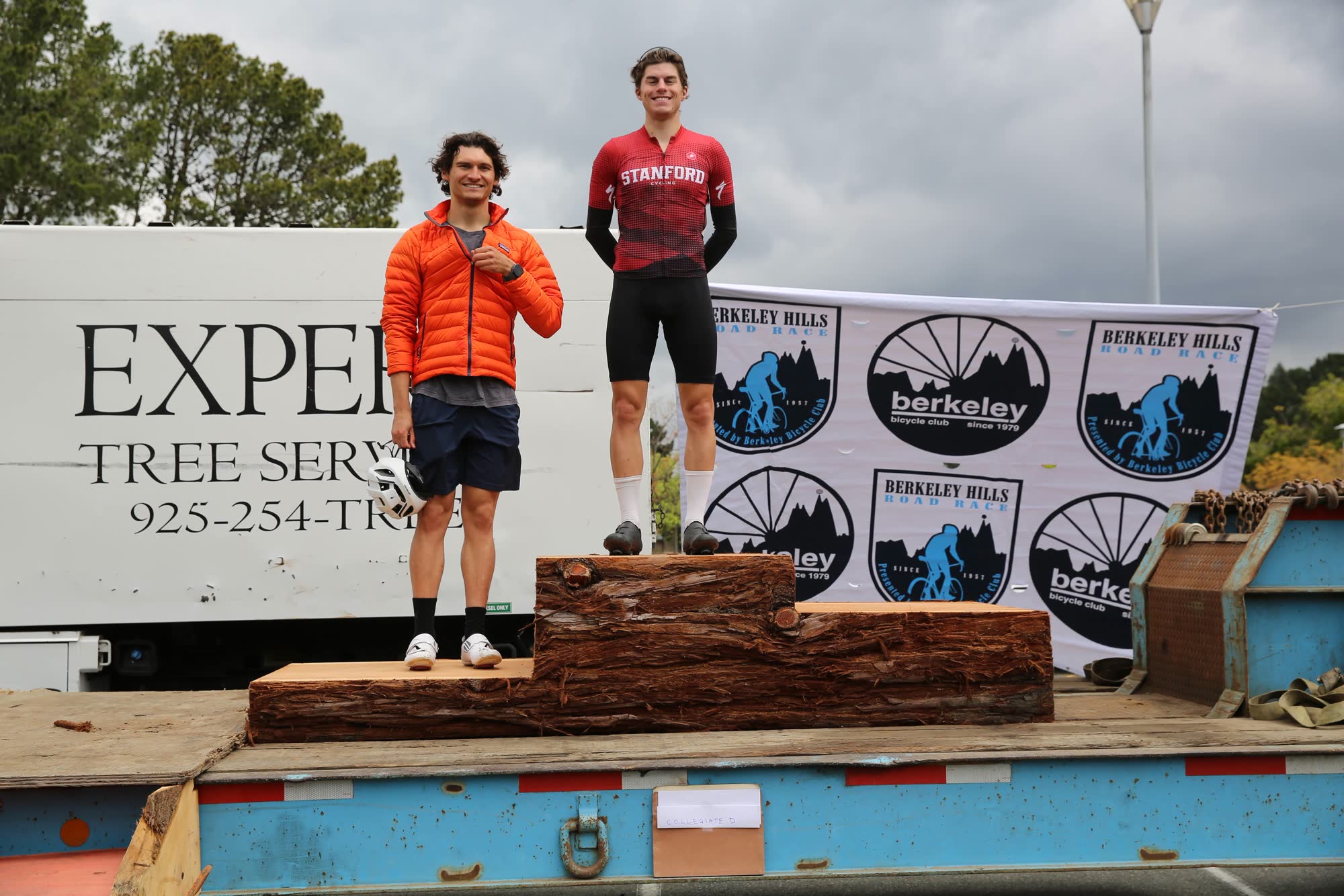 Two collegiate riders standing on a wooden podium in front of the Expert Tree Service truck and a Berkeley Hills Road Race banner