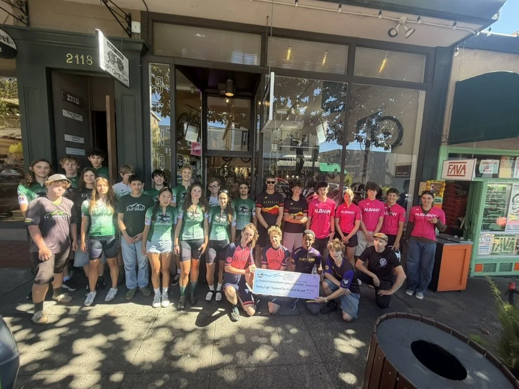 East Bay high school NICA team members posing outside a bike shop with a large check