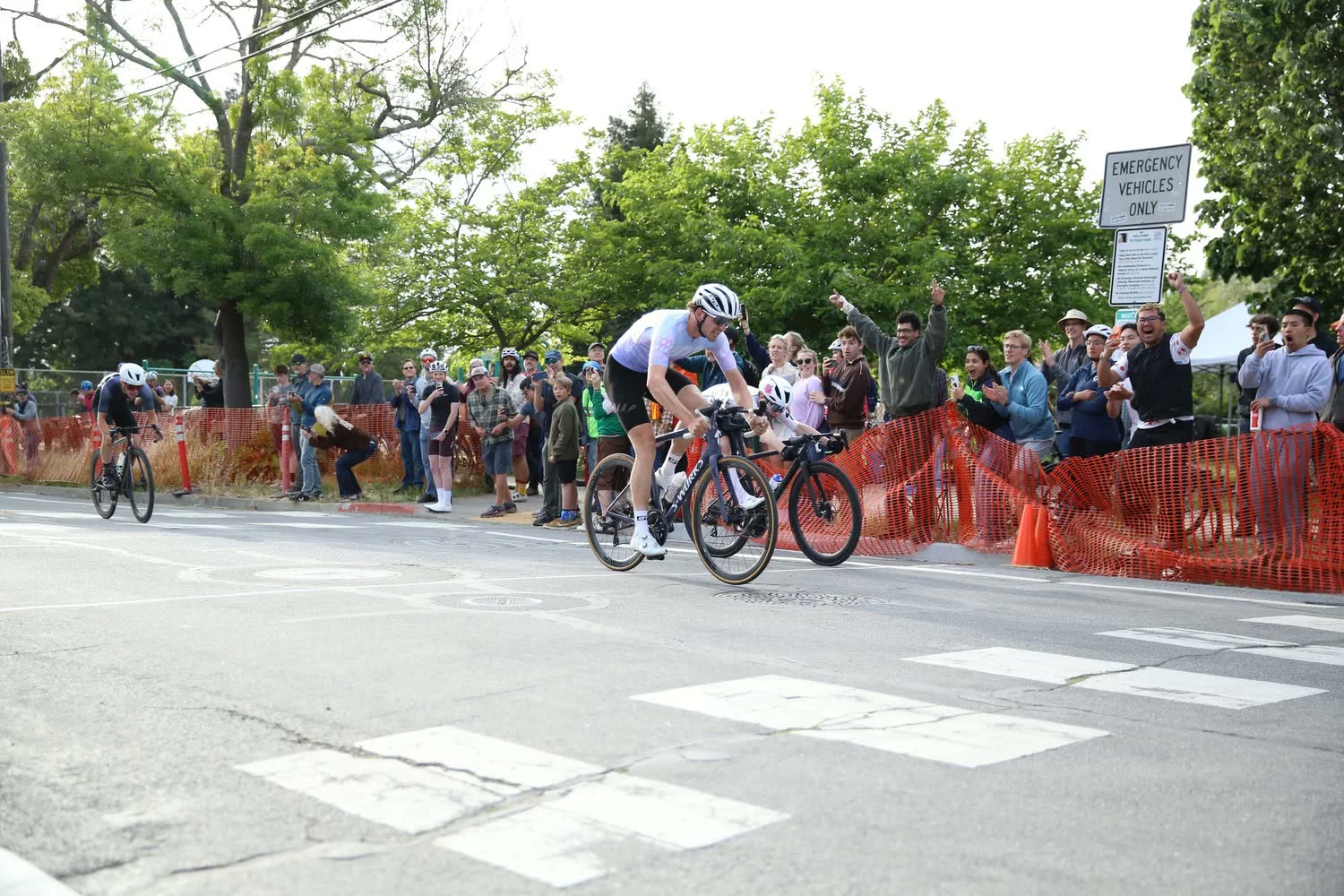 A rider in white and another in pink riding a corner past spectators behind orange fencing