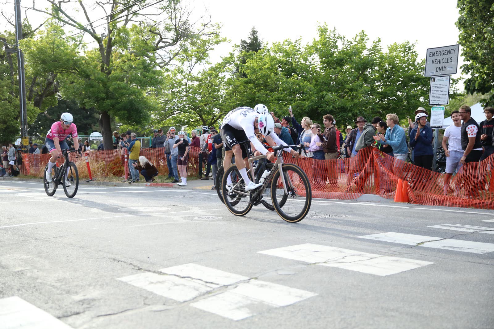 A rider in white chasing a rider in pink past spectators behind orange fencing at the Berkeley Streets Criterium