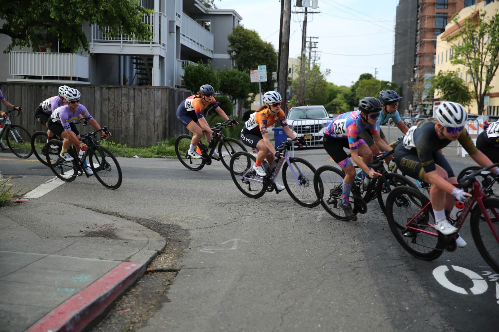 Riders in team kits leaning through a tight corner beside a red curb at the Berkeley Streets Criterium