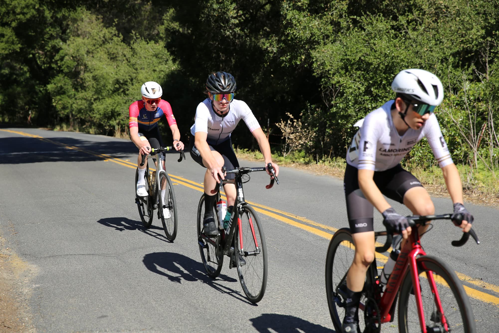 Three junior riders on a shaded road, the closest in a white kit