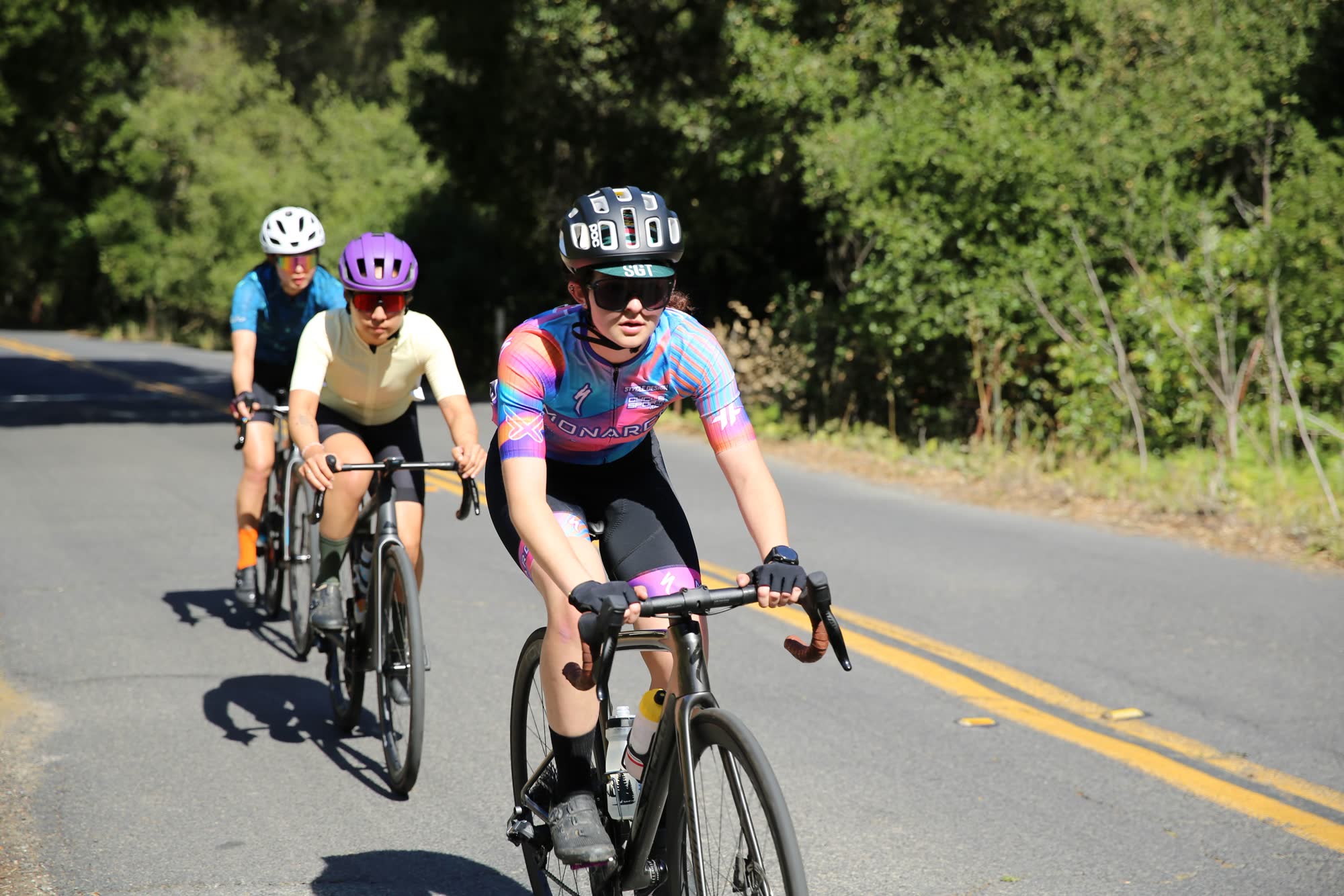 A junior rider in a bright pink, blue and orange kit leading a small group along a shaded road