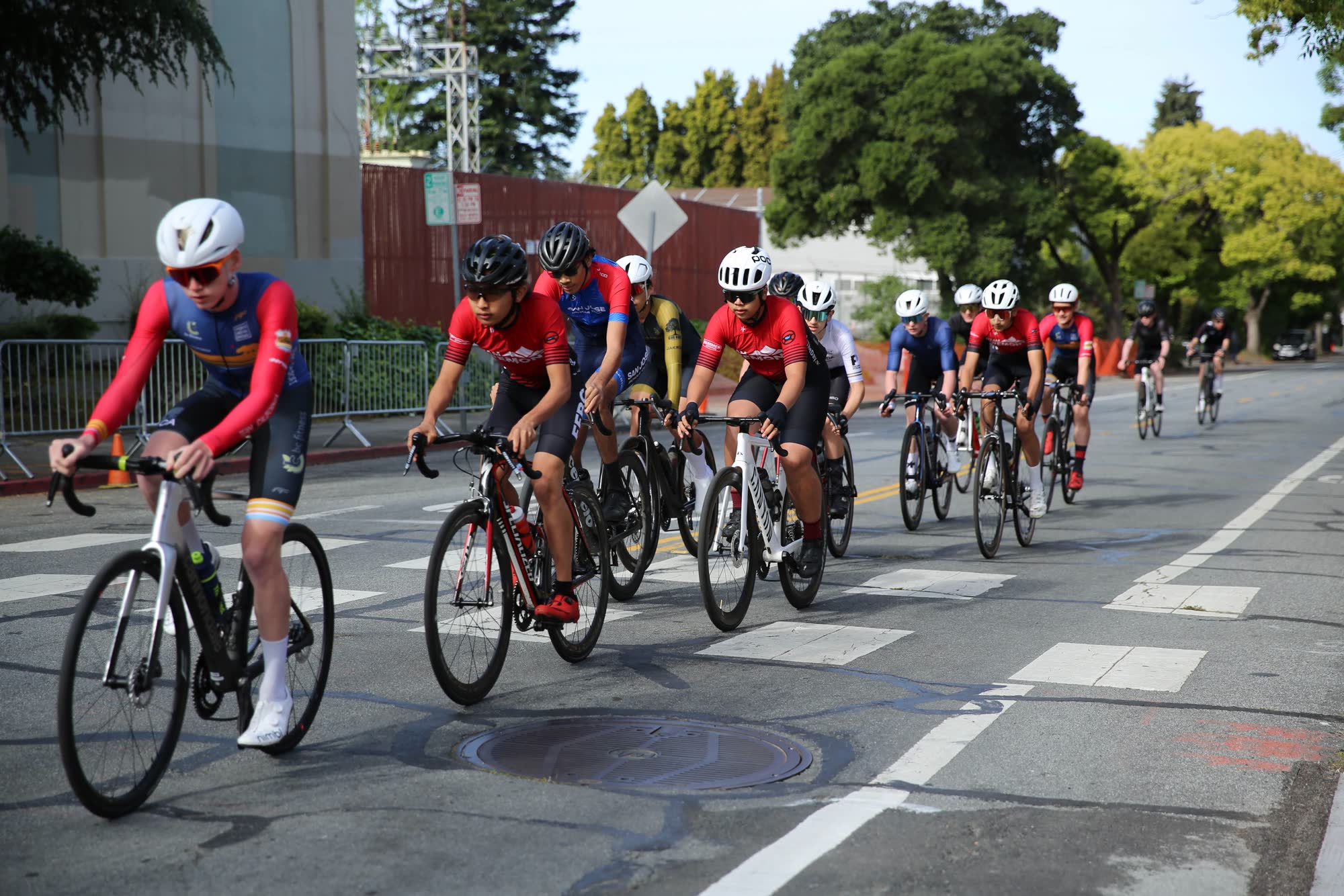 A line of junior riders in red and blue kits riding together down a street lined with barriers