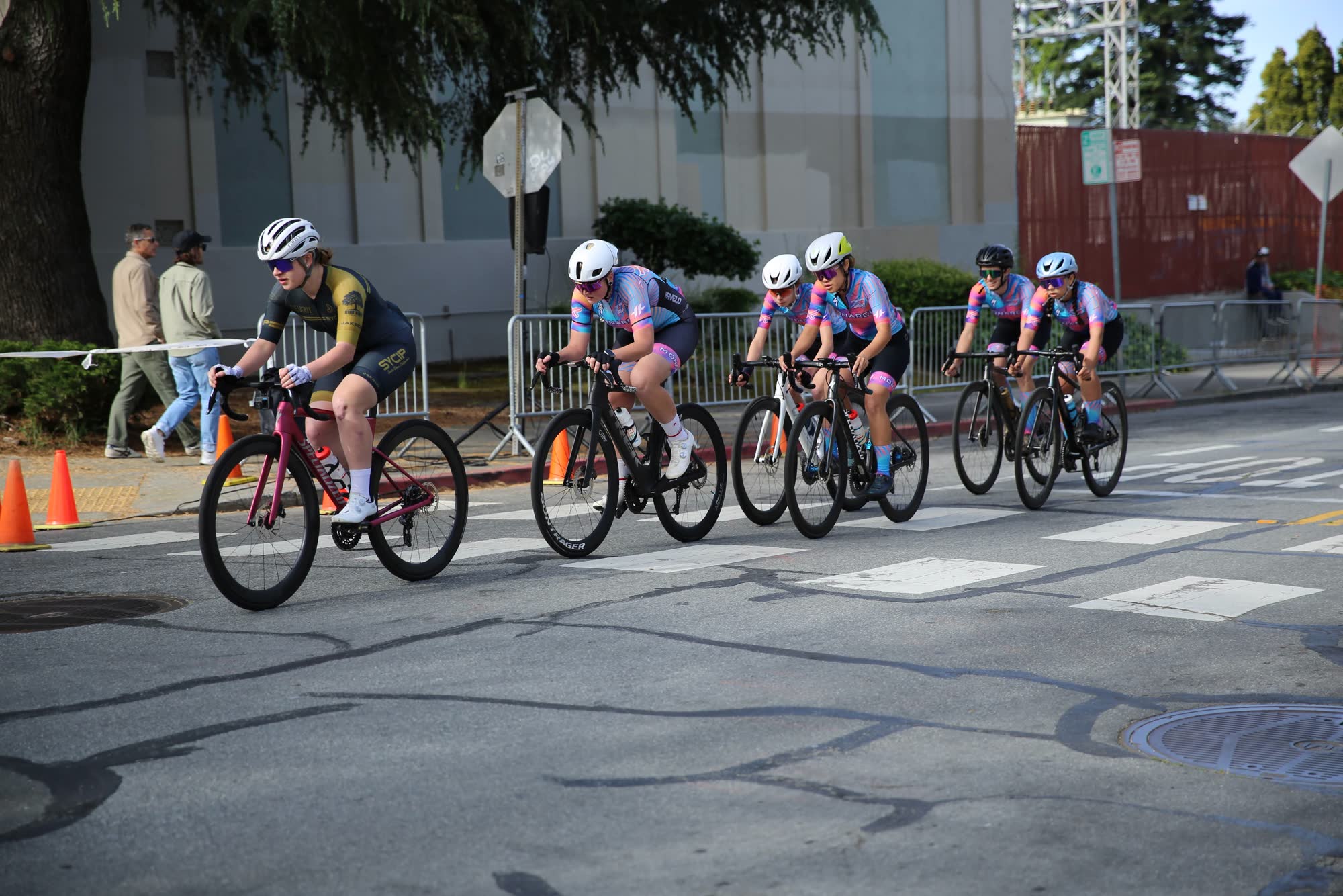 A group of junior riders in team kits riding past barriers on a downtown street