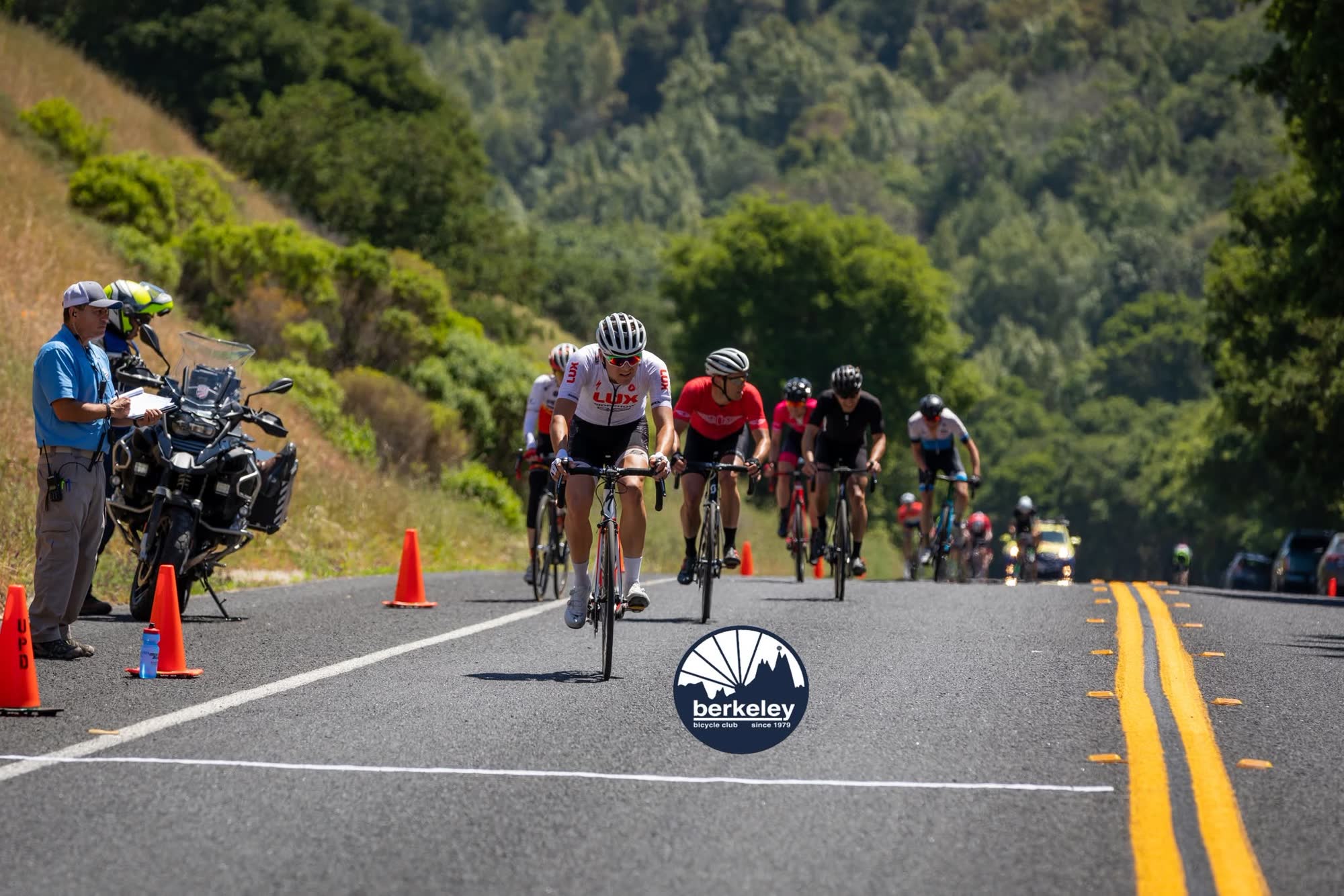 A group of riders approaching the finish line on a hillside road, with a race motorcycle and an official at the roadside