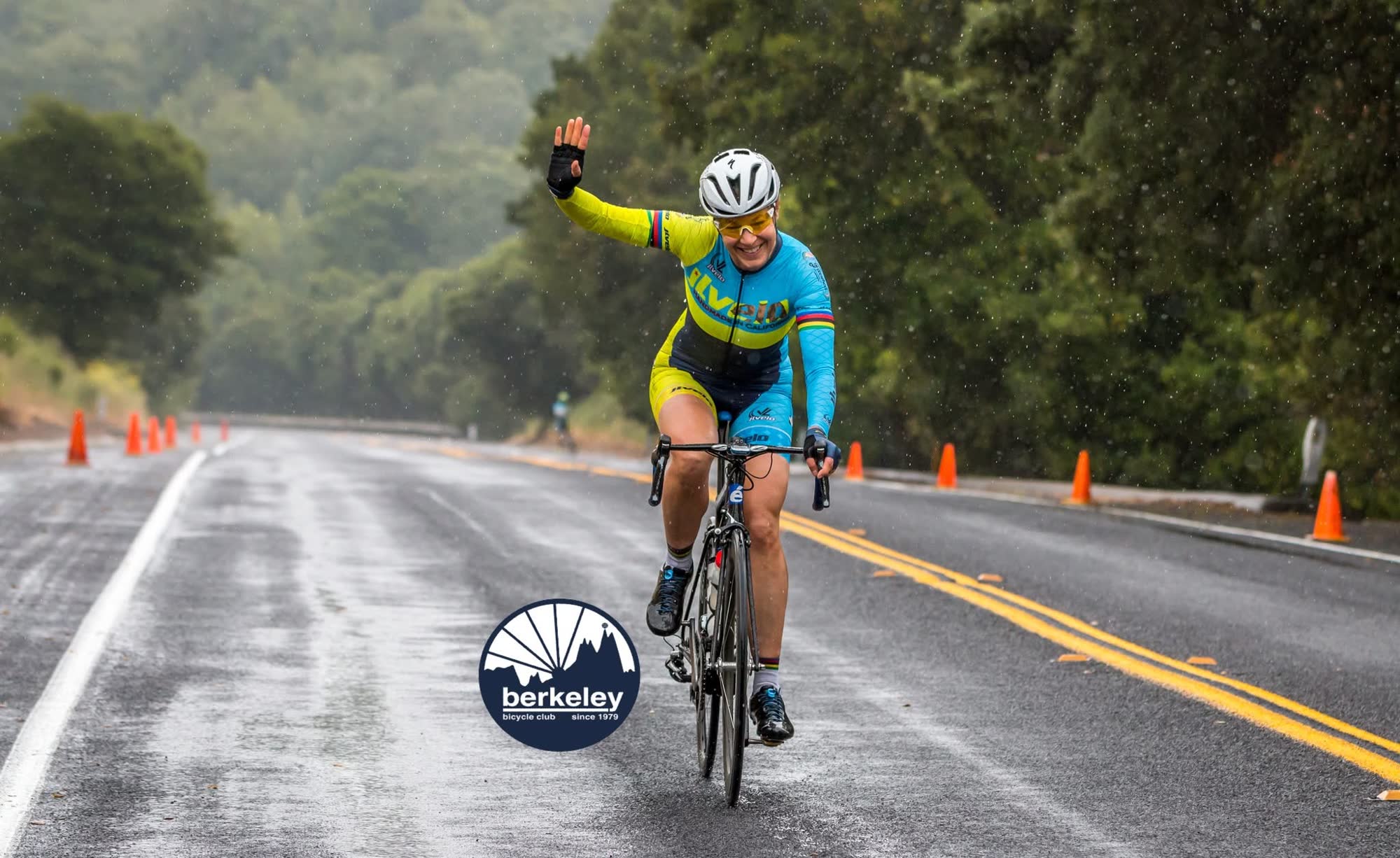 A rider in a bright yellow and blue kit raising one hand in celebration as they cross the line on a wet road
