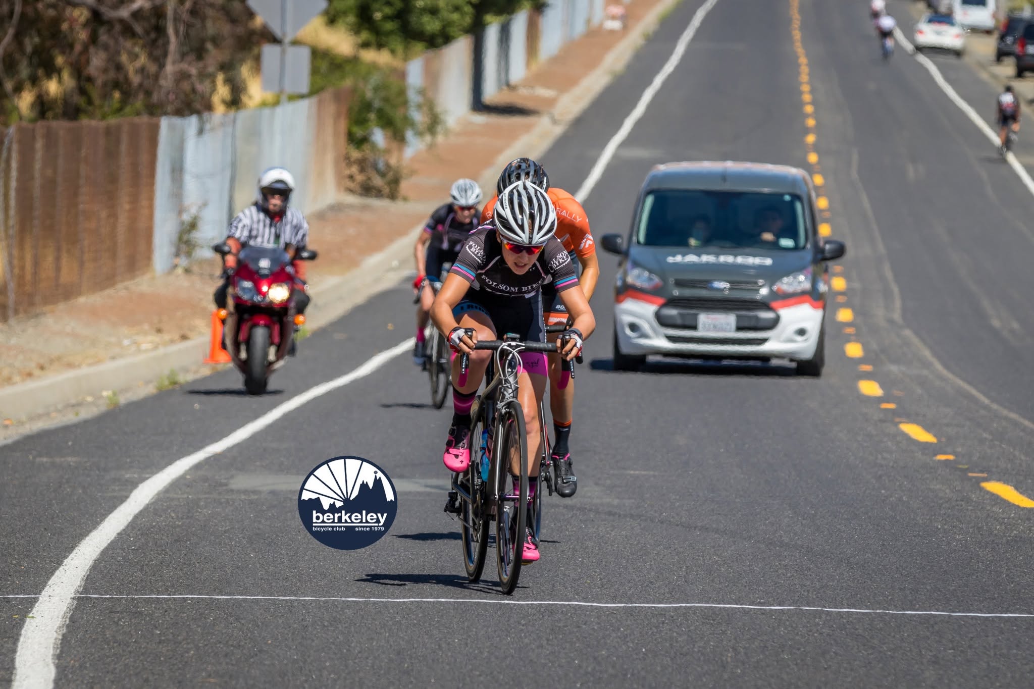 Riders sprinting toward the line with a race motorcycle and a support van behind them