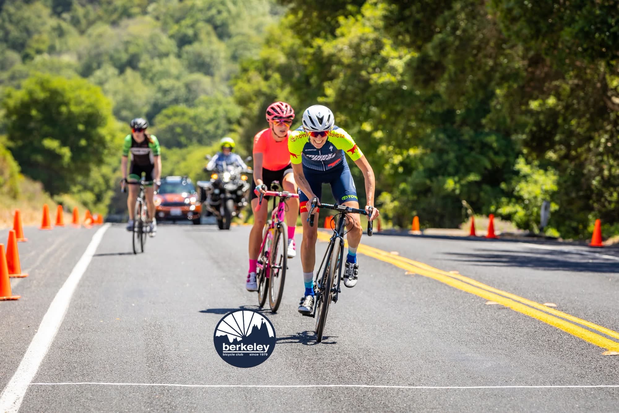 Two riders leading the chase to the finish along a road lined with orange cones, a race motorcycle following
