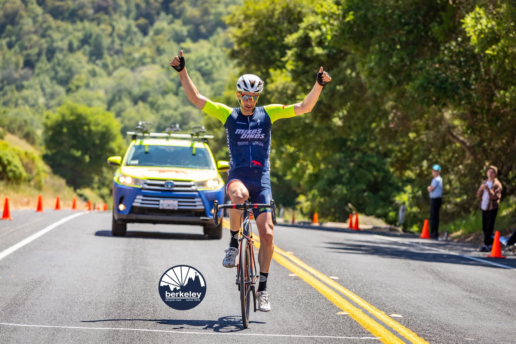A rider in a navy kit with lime sleeves crossing the line with both arms raised, a yellow support car behind them