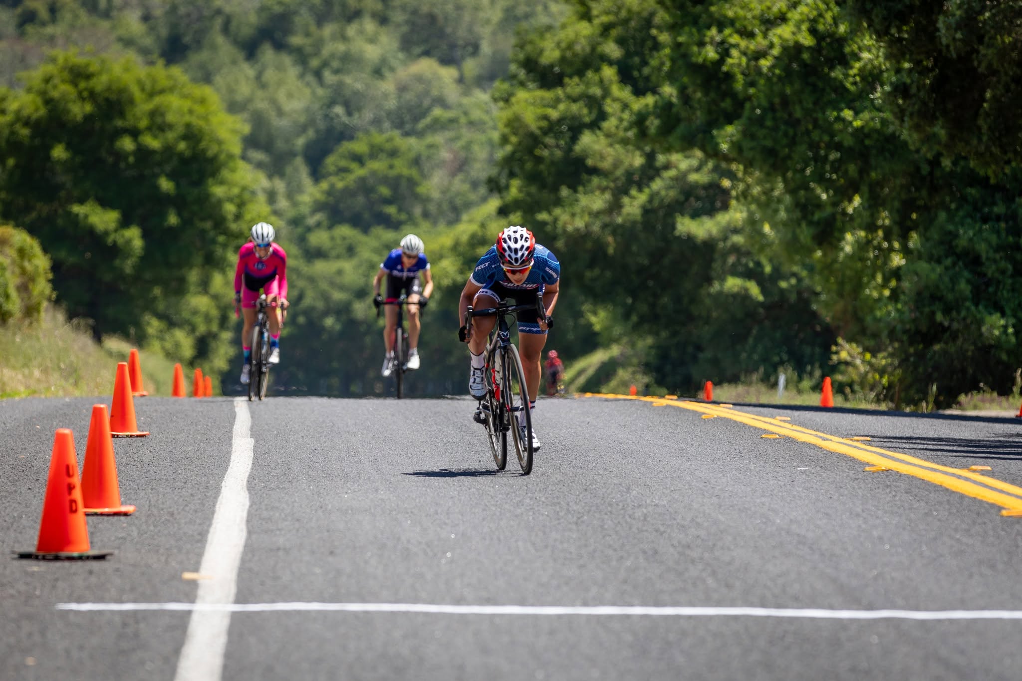 Three riders approaching the finish line along a road lined with orange cones