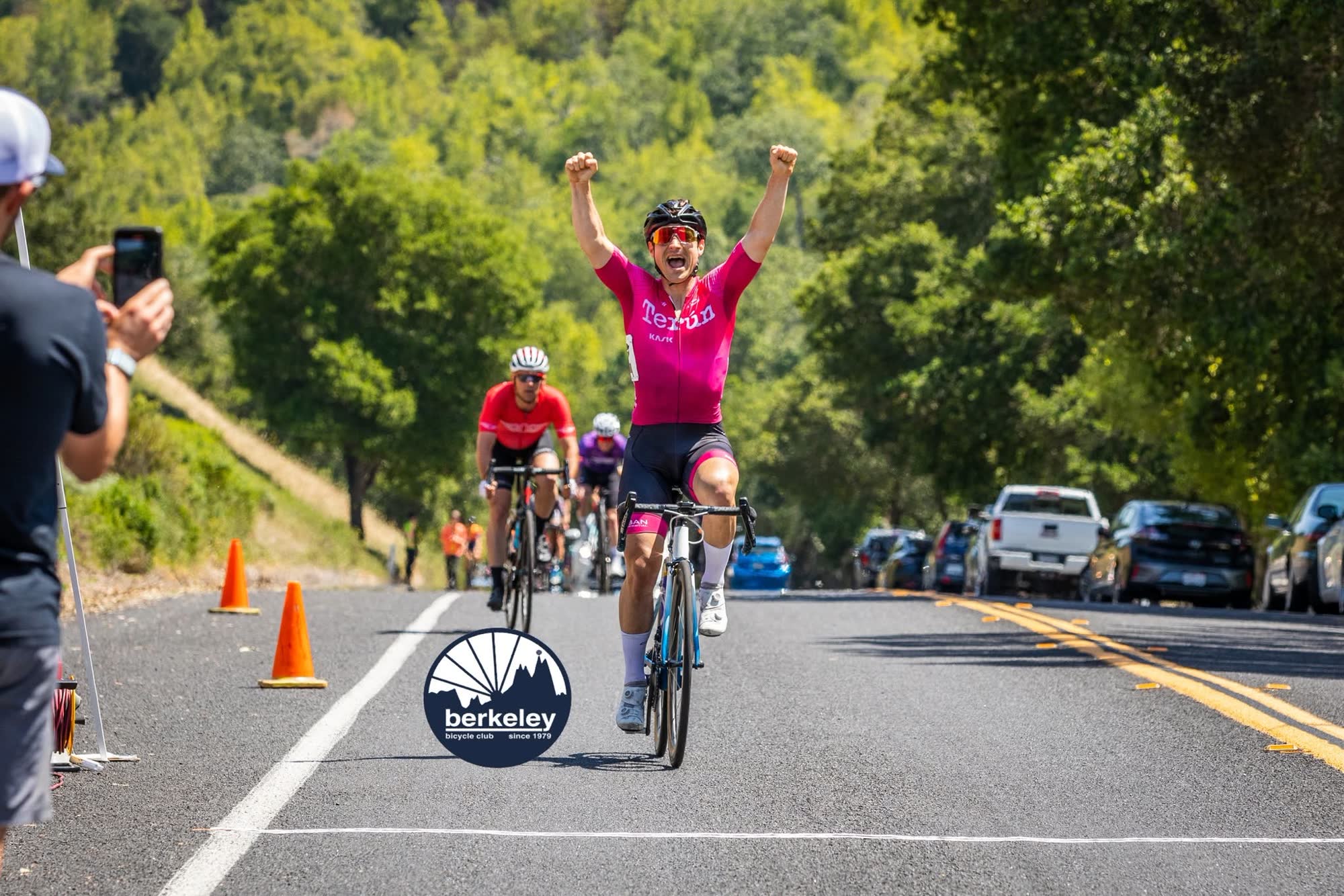 A rider in a pink kit raising both arms in celebration at the line while a spectator films on a phone