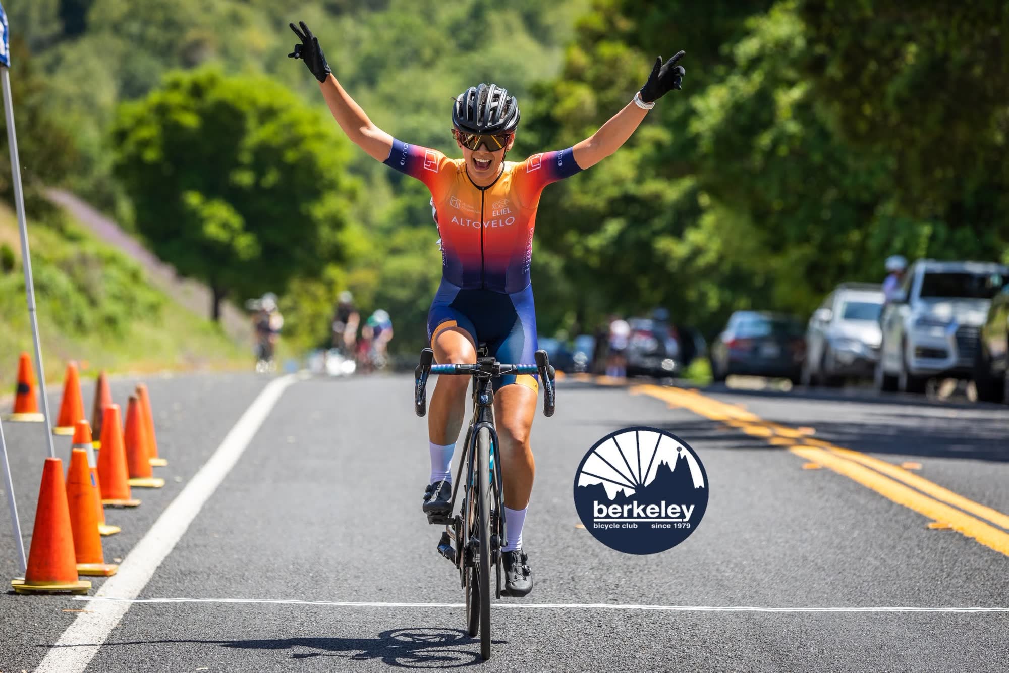 A rider in an orange and purple kit raising both arms as they cross the finish line