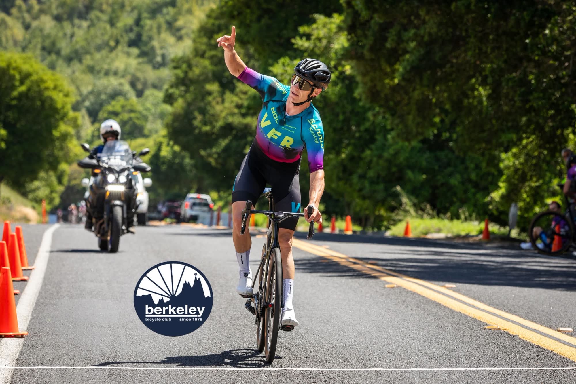 A rider in a teal, purple and pink kit raising one arm at the finish, with a race motorcycle behind