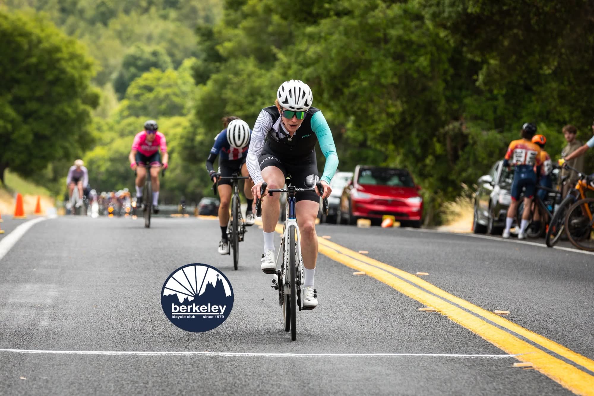 A rider in a black and white kit with teal sleeves leading a small group toward the line, with cars parked along the road