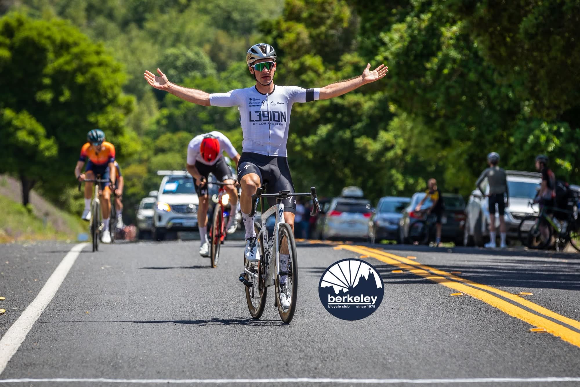 A rider in a white kit spreading their arms wide as they cross the line, with chasers behind