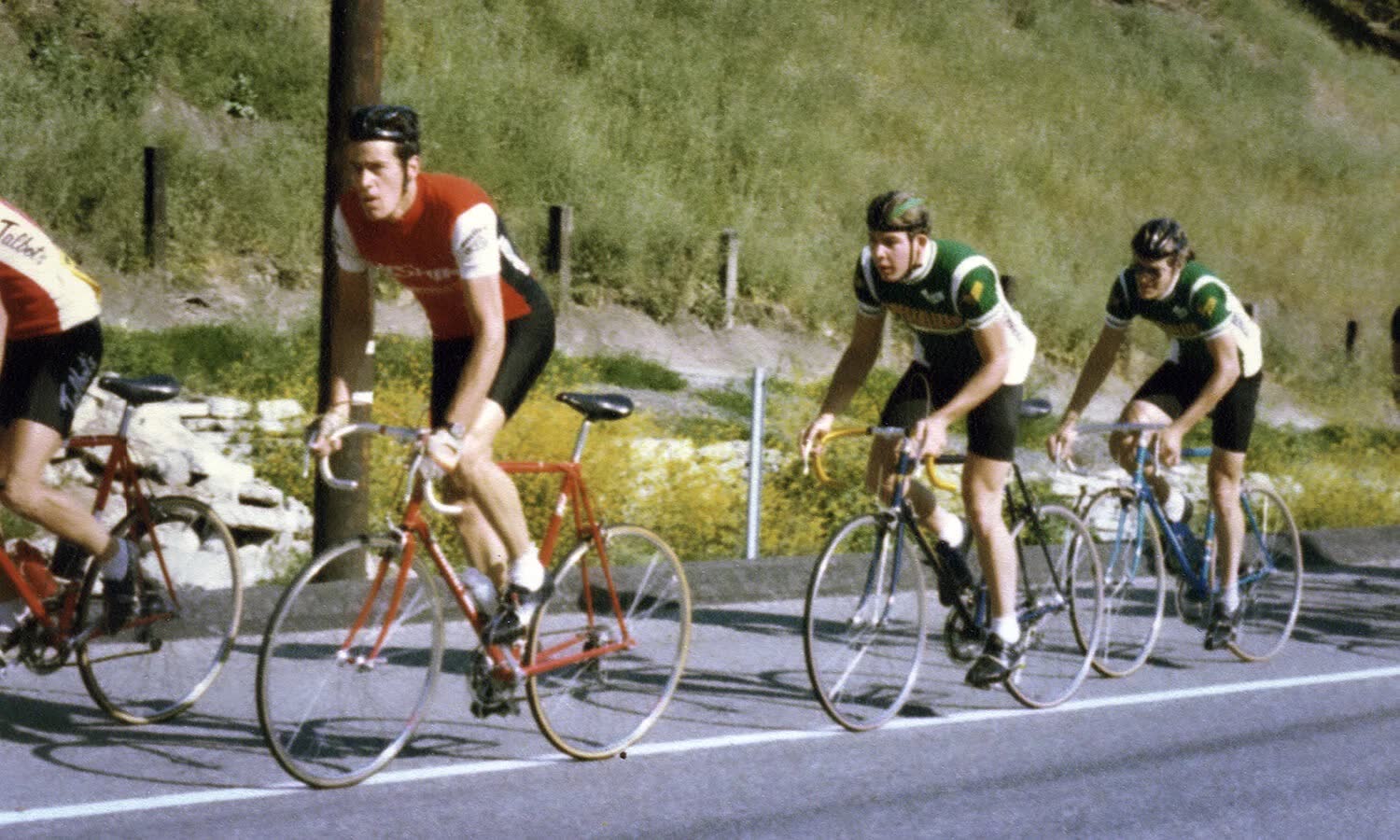 A small group of riders on a road below a grassy hillside