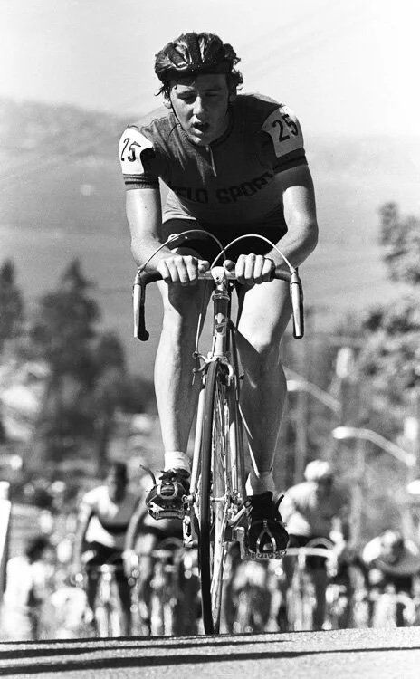 A rider in a Velo Sport jersey, number 25, climbing a road with the field behind