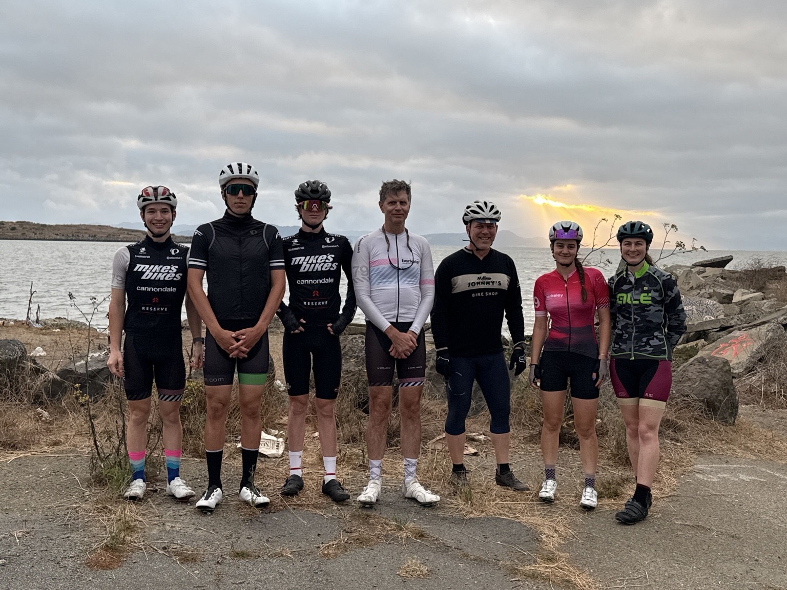 Junior and adult riders standing together on the bay shoreline before a ride, with light breaking through the clouds