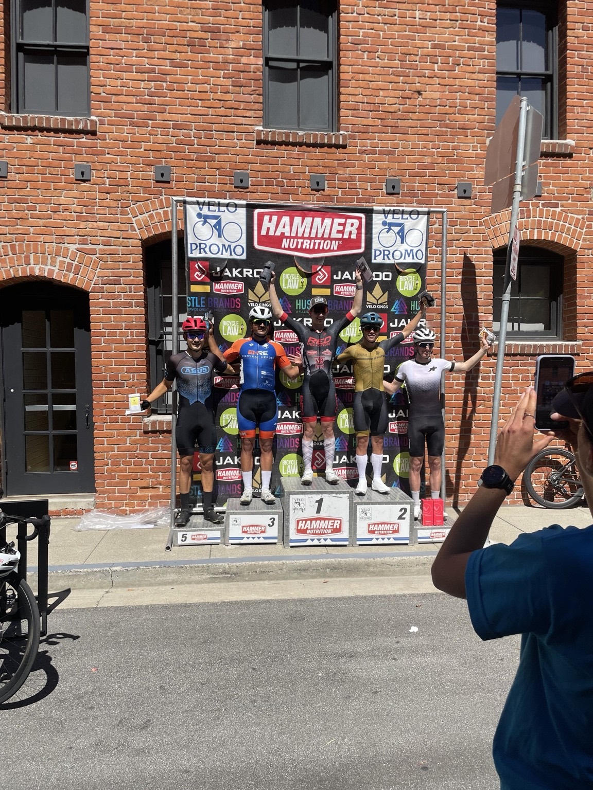 A rider in a Red Peloton kit with arms raised on the top step of a five-place podium in front of a brick building