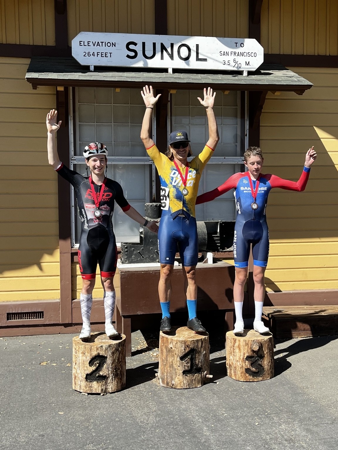 Three riders waving from wooden log podium steps in front of the Sunol train depot, second place in a Red Peloton kit
