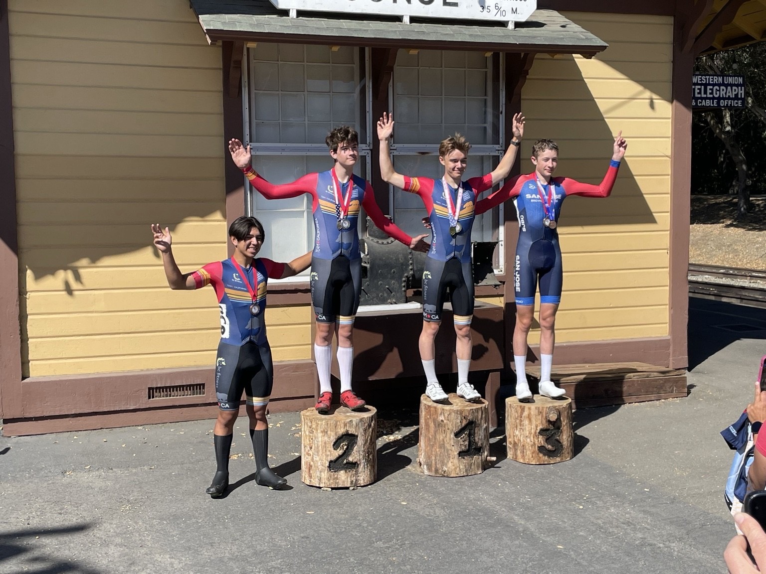 Young riders in Team California and San Jose Bicycle Club kits on the log podium at the Sunol train depot