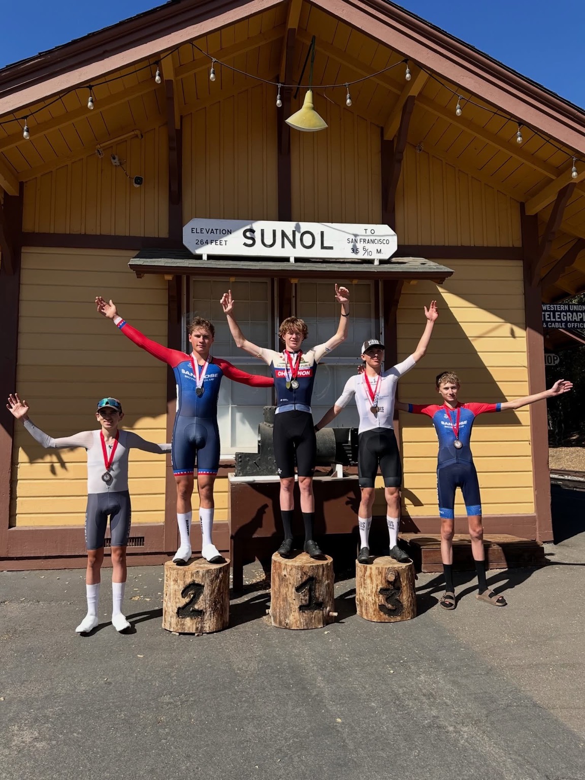 Five junior riders with medals celebrating on and beside the log podium under the Sunol station sign