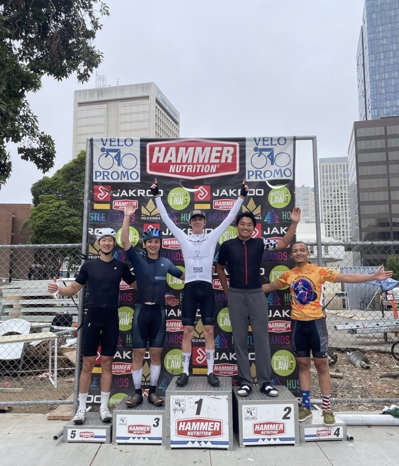 A rider in a white Sharp Bicycle kit on the top step of a five-place podium, arms raised, with city buildings behind