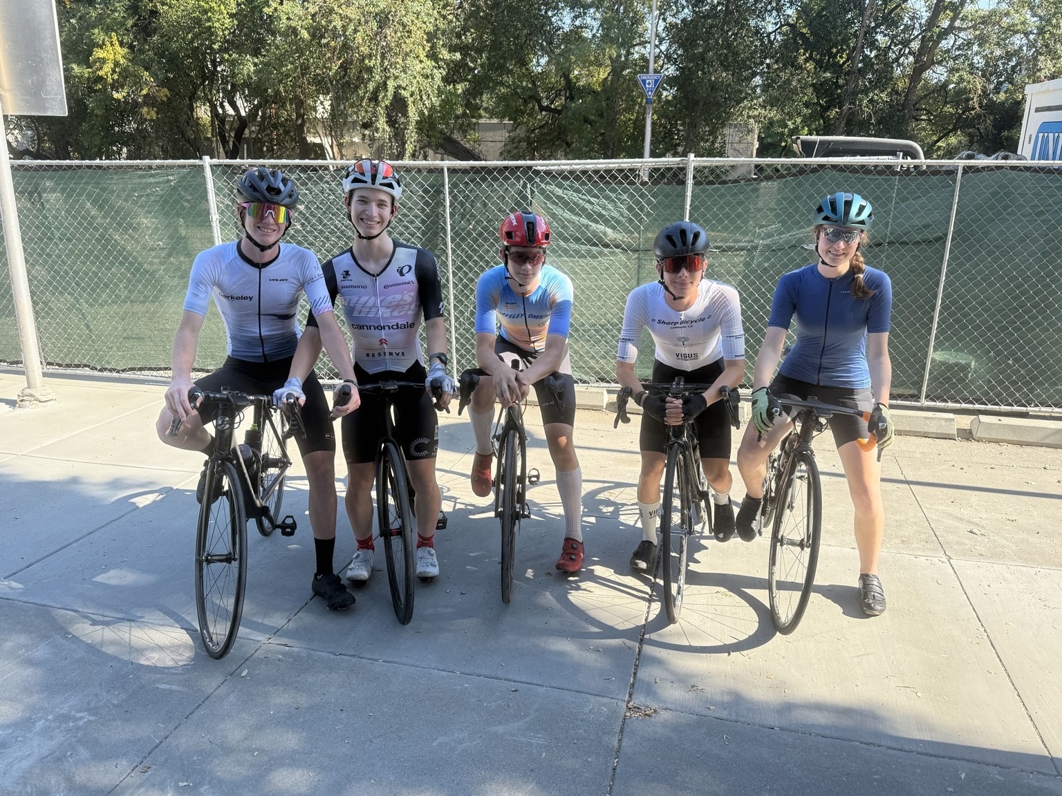 Five young riders in different team kits, including Berkeley, Mike's Bikes and Sharp Bicycle, leaning on their bikes together in front of a fence