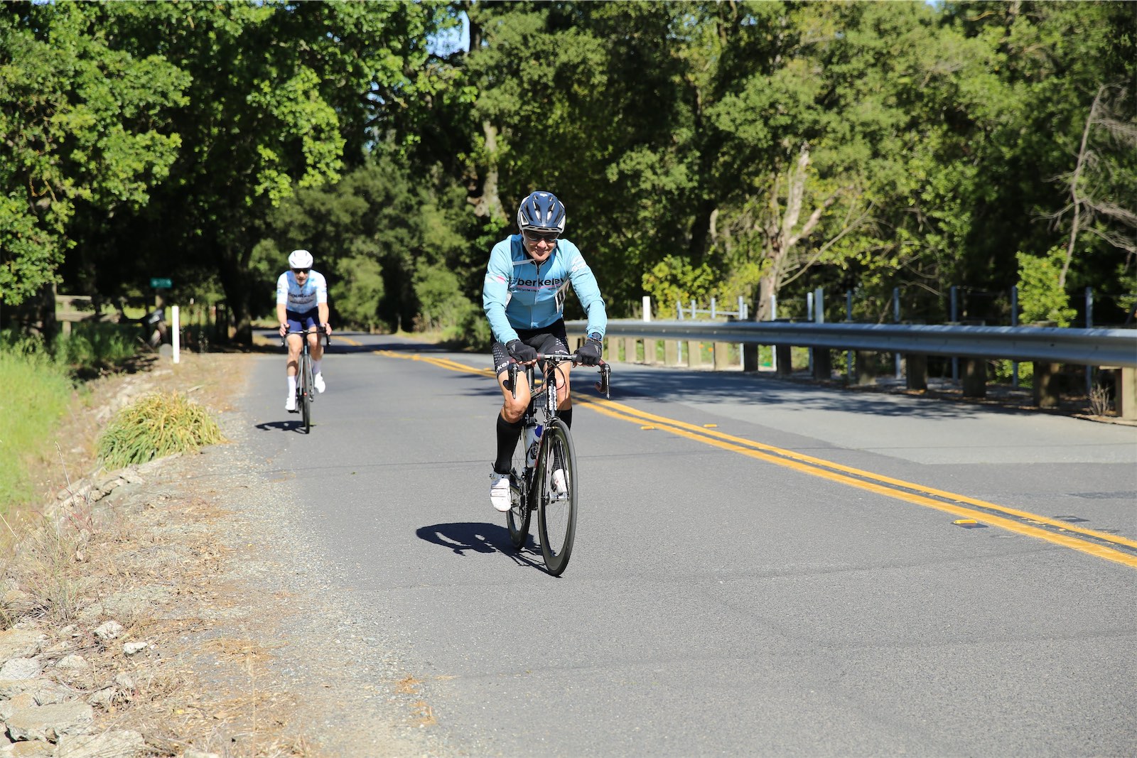 A Berkeley Bicycle Club rider in the club's light blue jersey climbs a sunny, tree-lined road at the 2026 Berkeley Hills Road Race, with another racer behind