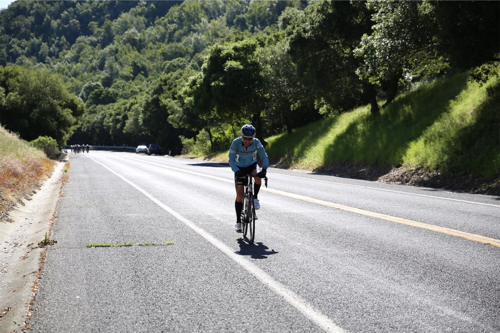 A Berkeley Bicycle Club rider in a light blue club jersey racing alone up a wide road through green hills at the 2026 Berkeley Hills Road Race