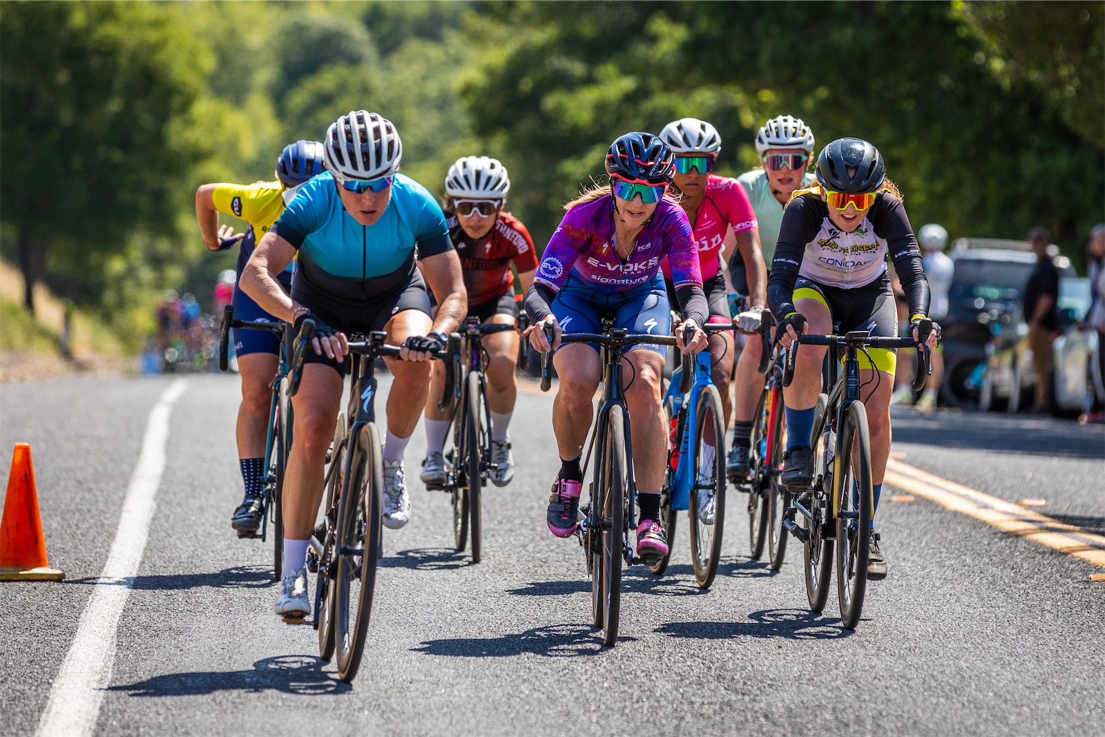 A pack of women racers on a sunny tree-lined road, led by riders in teal, purple E-Voke Racing and black-and-yellow kits, with more riders and spectators behind