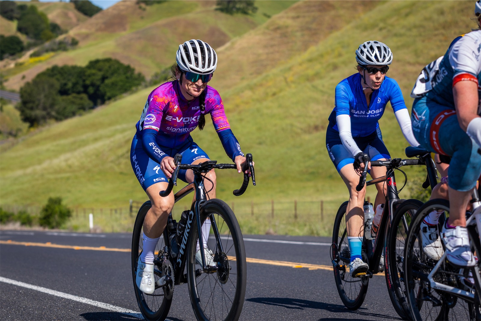 An E-Voke Racing rider in a purple and blue kit rides beside a San Jose Bicycle Club rider in blue, with green and golden hills behind them