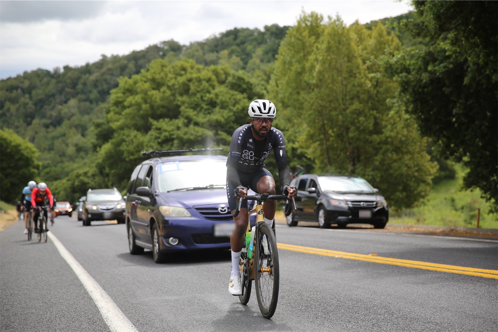 A Good Guys Racing rider in a black team kit and white helmet on a gold road bike, riding ahead of other racers and cars on a tree-lined road in the Berkeley hills