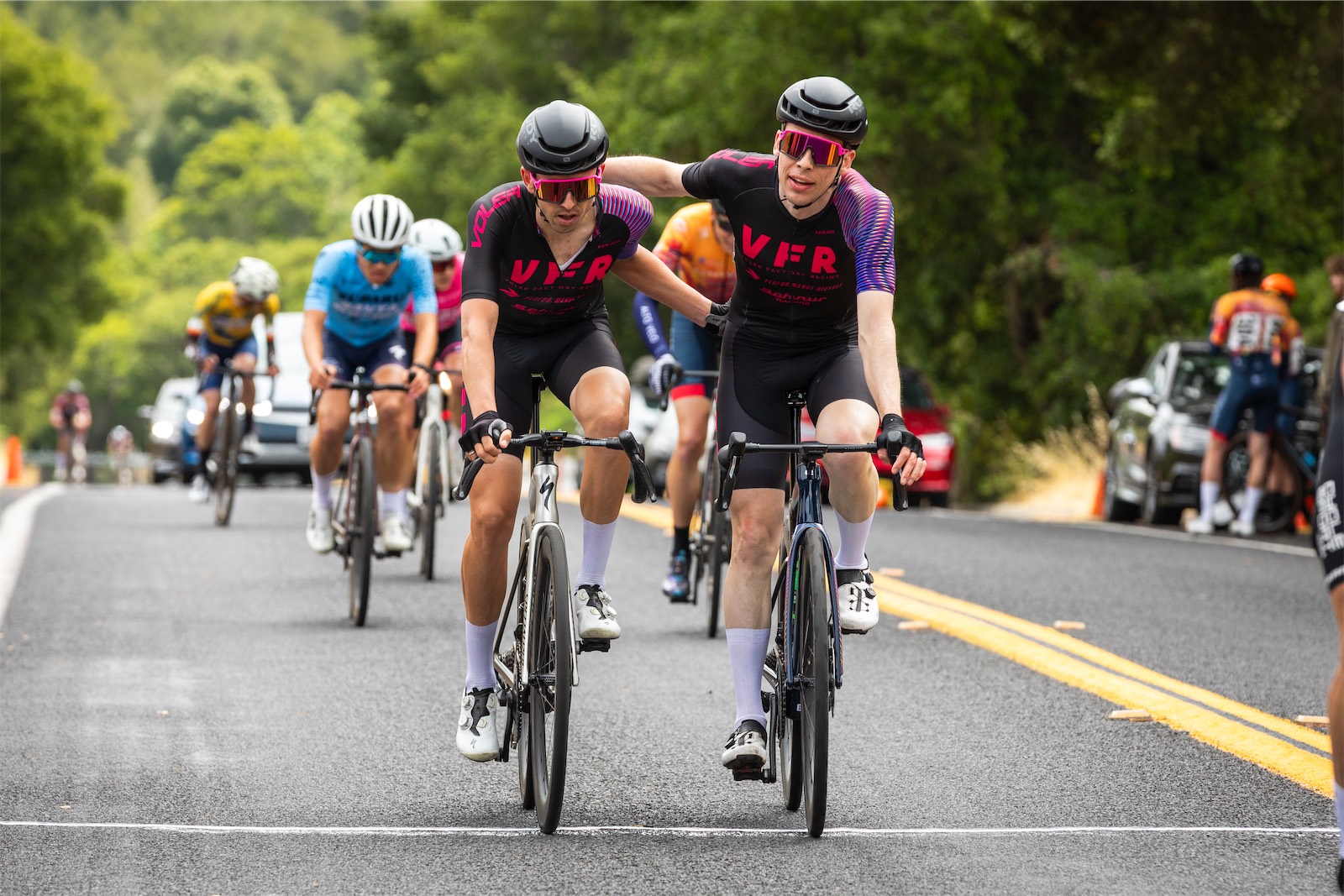 Two Voler Factory Racing riders in black and pink kits cross the finish line side by side, one with an arm around the other, as riders in other team kits follow on a tree-lined road