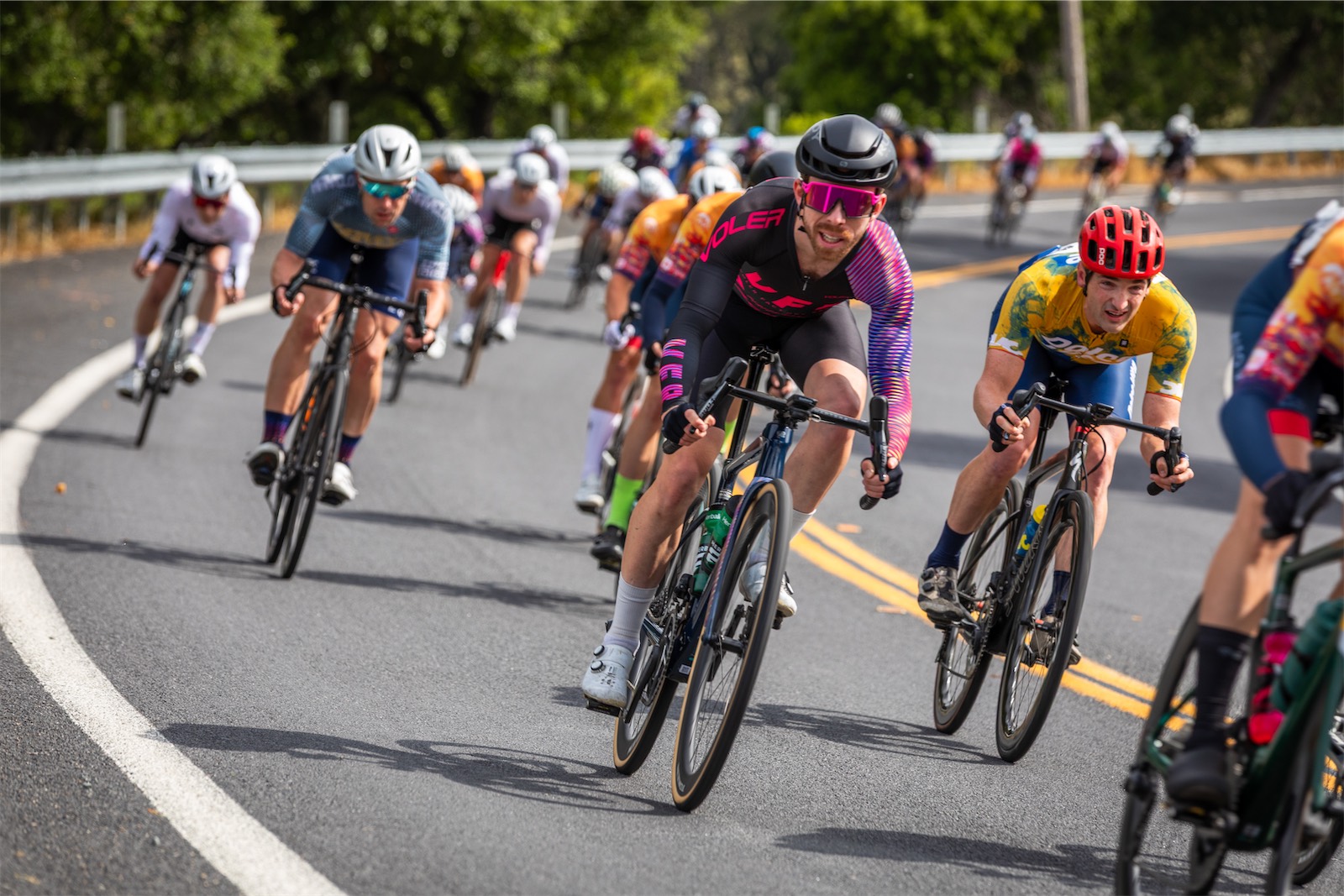 A Voler Factory Racing rider in a black and pink skinsuit leads a pack of riders through a curve on a tree-lined road